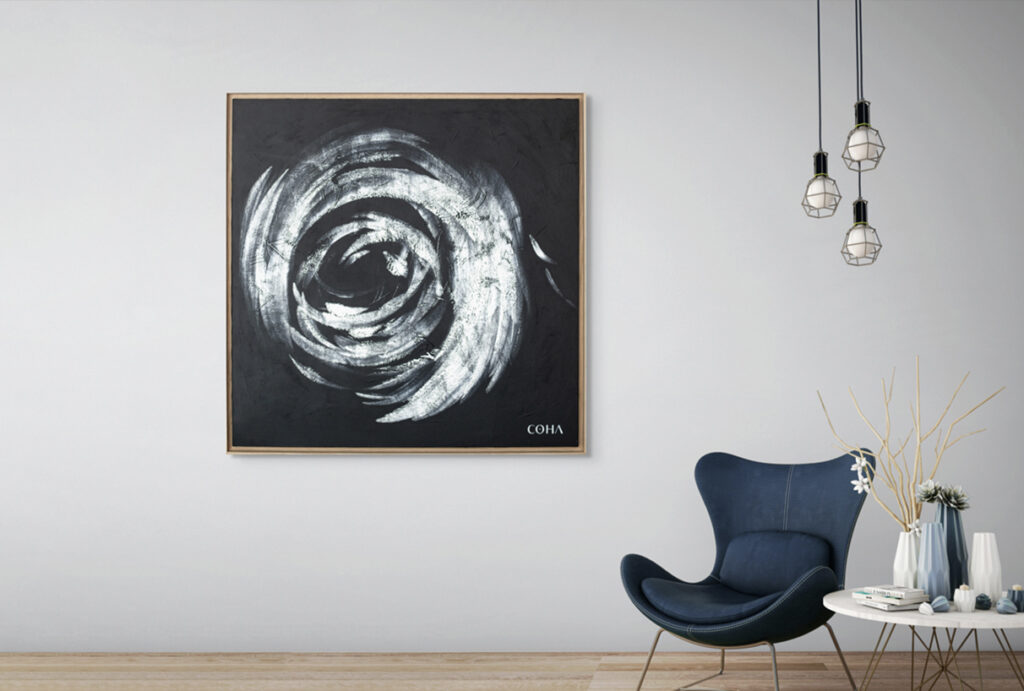 Tableau moderne COHA accroché à un mur blanc, représentant un cercle abstrait en noir et blanc avec des coups de pinceau texturés. Le cadre est en bois clair, et le bas du tableau comporte la signature 'COHA'. À droite, un fauteuil bleu design est placé près d'une table basse blanche ornée de décorations minimalistes et d'une branche sèche. Trois suspensions lumineuses en métal noir complètent l'espace élégant et épuré.