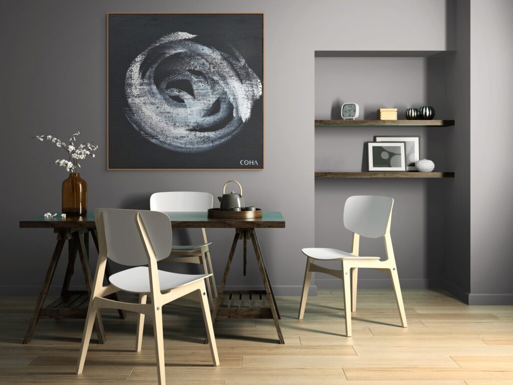 Cette image montre un espace à manger moderne et chaleureux. L'œuvre d'art au mur, signée "COHA," est une composition abstraite circulaire, réalisée dans des nuances de blanc et de gris sur un fond noir, apportant une touche artistique et contemporaine à la pièce.

Le mobilier inclut une table en bois rustique soutenue par des tréteaux, entourée de chaises design en bois clair avec des assises blanches, qui allient modernité et simplicité. Le décor est complété par une petite étagère murale avec des objets décoratifs minimalistes, ainsi qu'un vase en verre ambré contenant des branches fleuries. Les tons gris des murs et le parquet en bois clair créent une ambiance douce et accueillante.