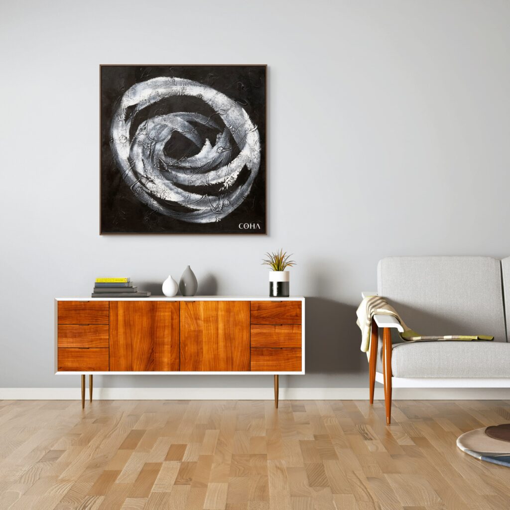 Cette image présente une pièce moderne et minimaliste avec un sol en bois clair, un buffet au design rétro en bois et blanc, un fauteuil gris clair avec des pieds en bois et une couverture posée dessus. Un tableau abstrait, dominé par des tons noirs et blancs et signé "COHA", est accroché au mur gris au-dessus du buffet. Sur le buffet, des objets décoratifs tels qu'une plante, des livres et des vases ajoutent une touche d'élégance. L'ambiance est chaleureuse et contemporaine.