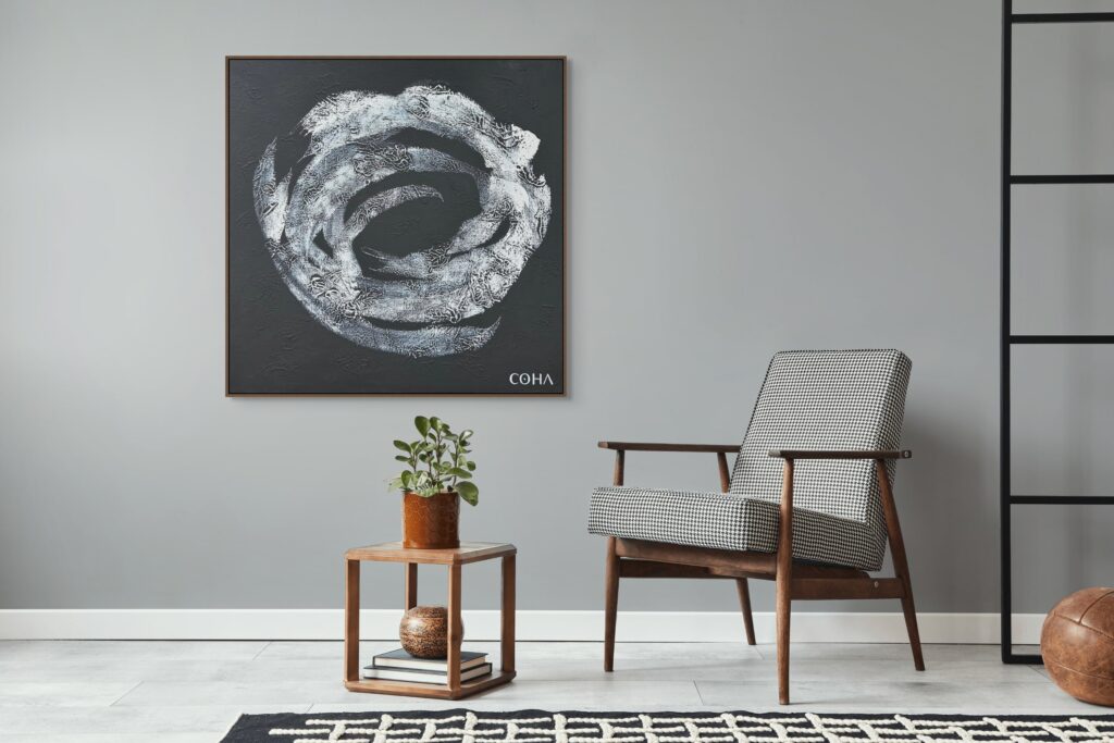 Cette image montre un intérieur minimaliste et chic avec une palette de couleurs neutres. Un tableau abstrait en noir et blanc signé "COHA" est accroché au mur gris clair, apportant une touche artistique. Un fauteuil au style rétro, avec une structure en bois et un revêtement en tissu à motif pied-de-poule, est disposé à côté d'une petite table en bois. Sur la table, une plante verte en pot et quelques livres ajoutent une touche naturelle et décorative. Un tapis noir et blanc à motif géométrique recouvre une partie du sol en pierre claire, renforçant le style moderne et épuré de la pièce.